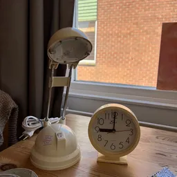 Een witte bureaulamp en een beige wandklok op een houten tafel bij een raam