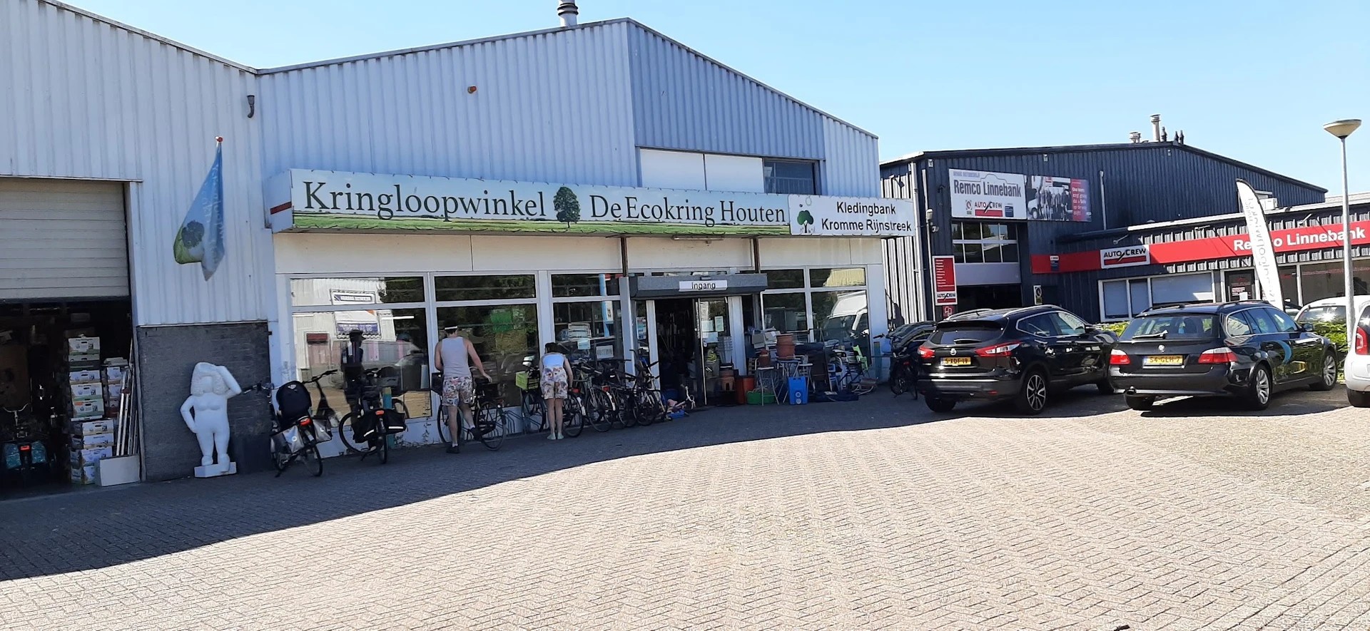 Gevel van kringloopwinkel met naam op gevel, ingang en fietsen ervoor, plavuizen plein