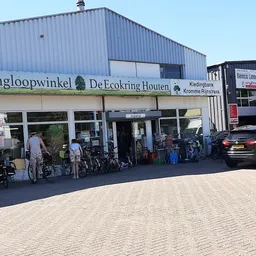Gevel van kringloopwinkel met naam op gevel, ingang en fietsen ervoor, plavuizen plein