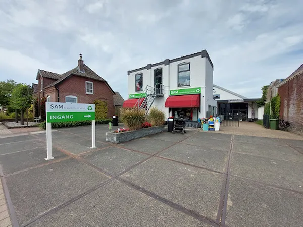 Voorgevel van kringloopwinkel met groene SAM-uithangbord en ingang, bakstenen gebouw links en wit-rood pand rechts