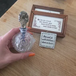 Close-up van een decoratieve glazen flesje met zilveren dop in de hand, met achtergrond van houten memobordjes met Nederlands