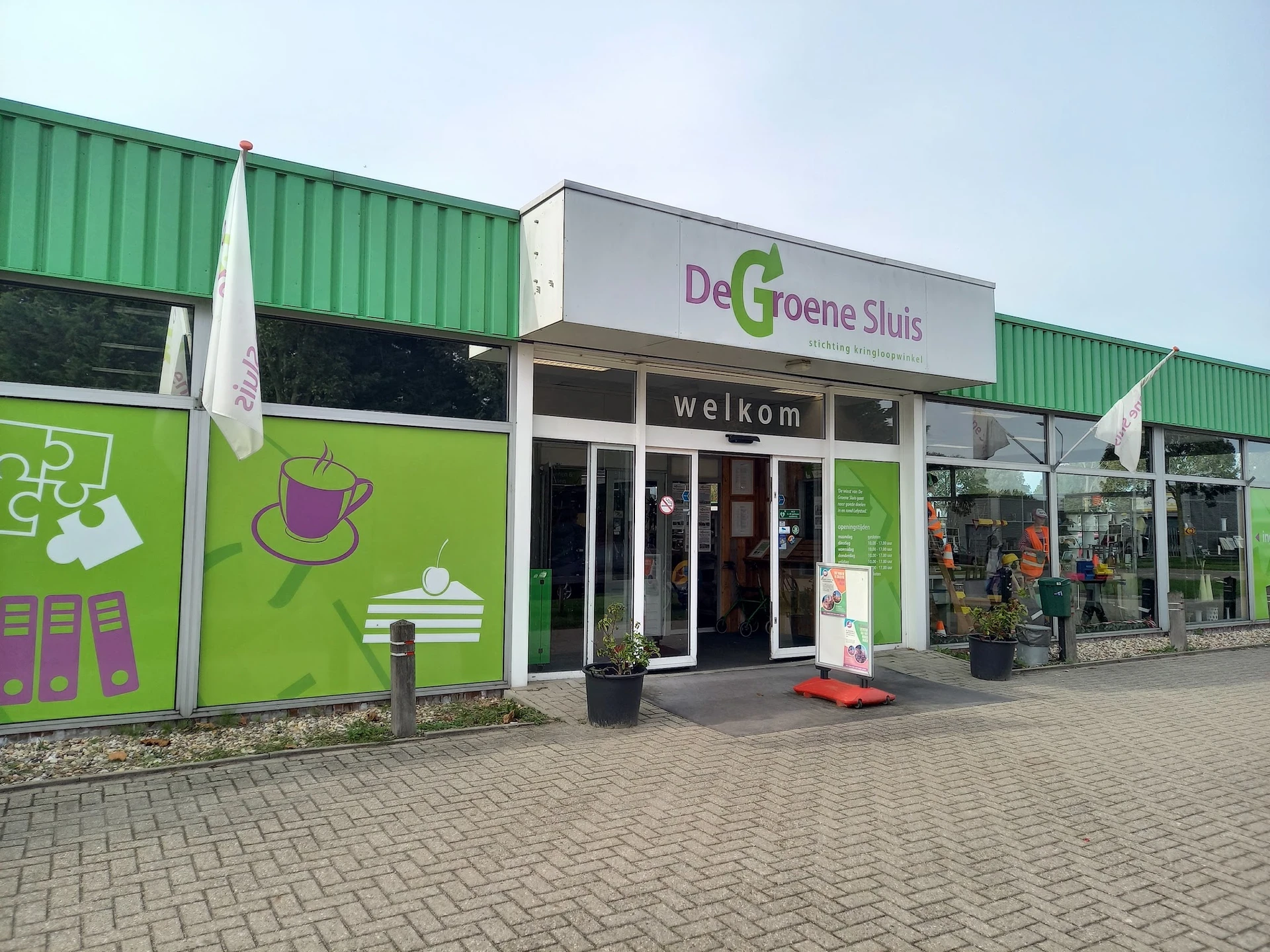 Groene gevel van kringloopwinkel met welkomstbord en glazen deuren aan de voorkant