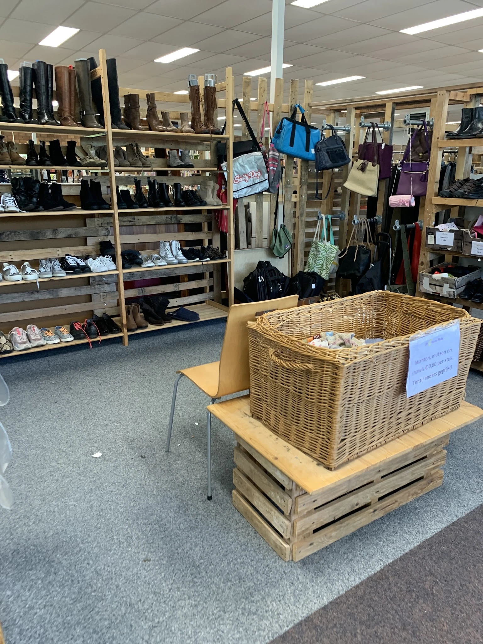 Grote rieten mand vol met tweedehands items op houten platform in kringloopwinkel, met stellingen en kleding op de achtergron