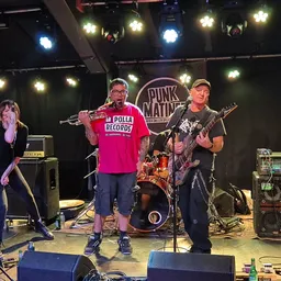 Band speelt op een podium met verlichting en 'Punk Mains' logo op de achtergrond