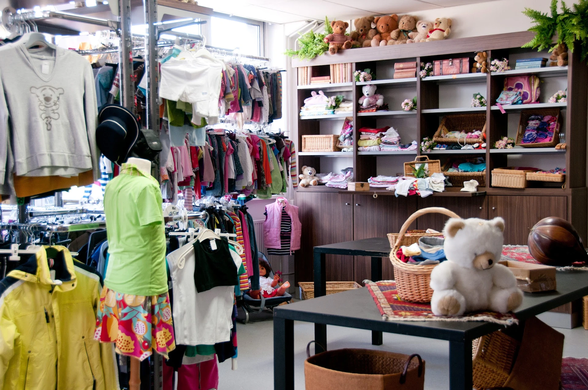 Binnenkant kringloopwinkel met kledingrekken, stellingen met huishoudartikelen en speelgoed, knuffeldieren op tafel