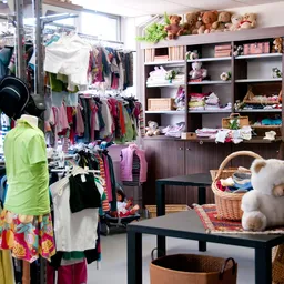 Binnenkant kringloopwinkel met kledingrekken, stellingen met huishoudartikelen en speelgoed, knuffeldieren op tafel