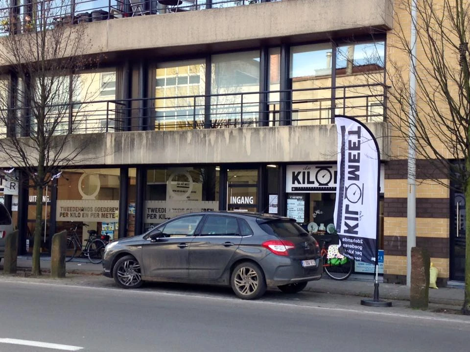 Gevel van kringloopwinkel De Kilomeet met ingang, grote ramen en blauwe vlaggenstandaard