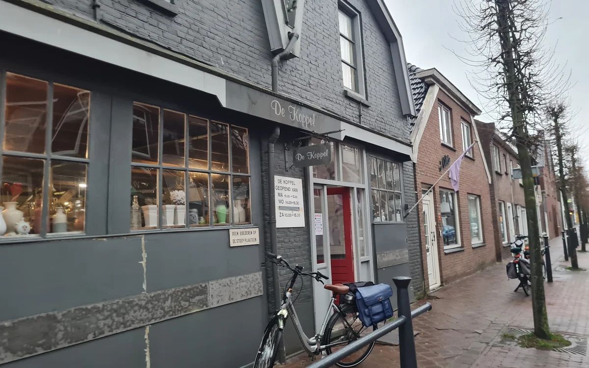 Straatgevel van kringloopwinkel met donkergroene pui, ramen met etalage, rode deur en fiets ernaast