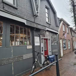 Straatgevel van kringloopwinkel met grijze pui, ramen en rode deur, fietsen geparkeerd ervoor