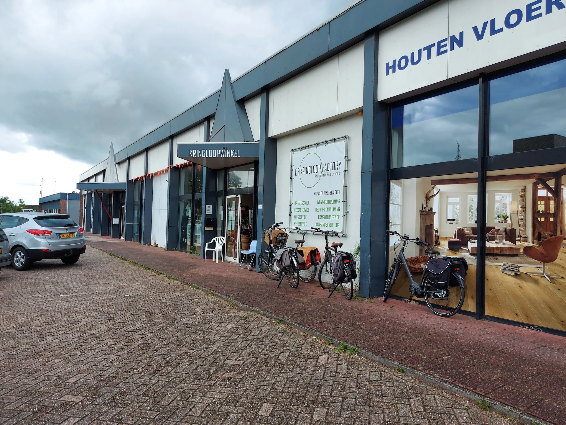 Voorgevel van kringloopwinkel met donkerblauwe lijsten, grote ramen, winkelnaam op gevel, fietsen voor ingang, parkeerplek li