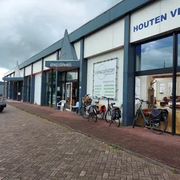 Voorgevel van kringloopwinkel met donkerblauwe lijsten, grote ramen, winkelnaam op gevel, fietsen voor ingang, parkeerplek li