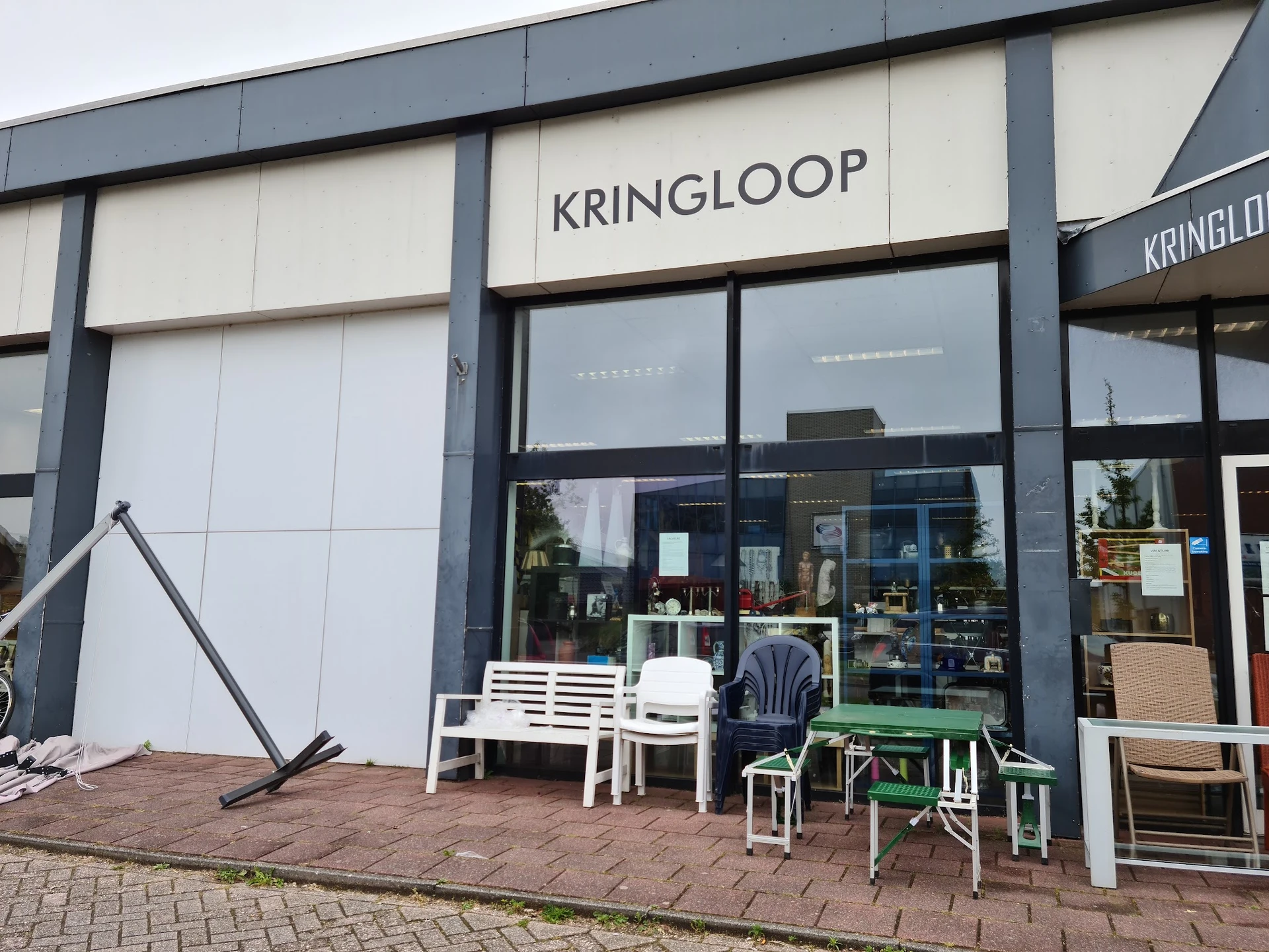 Gevel van kringloopwinkel met witte en grijze panelen, grote ramen, zitgroep buiten op terras