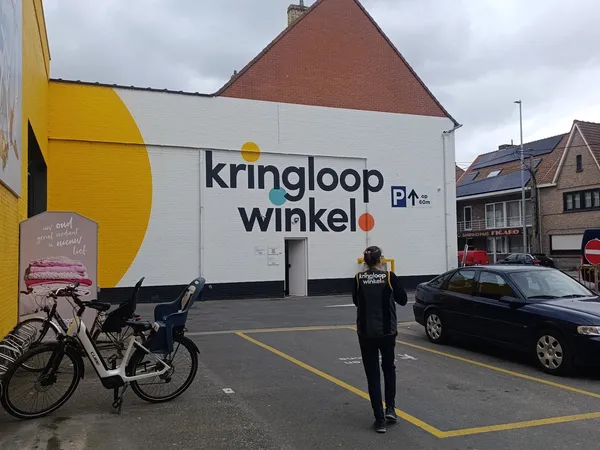 Witte gevel van kringloopwinkel met geel accent en grote letters, parkeerplaats met fietsen en auto's ervoor