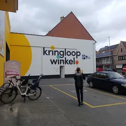 Witte gevel van kringloopwinkel met geel accent en grote letters, parkeerplaats met fietsen en auto's ervoor