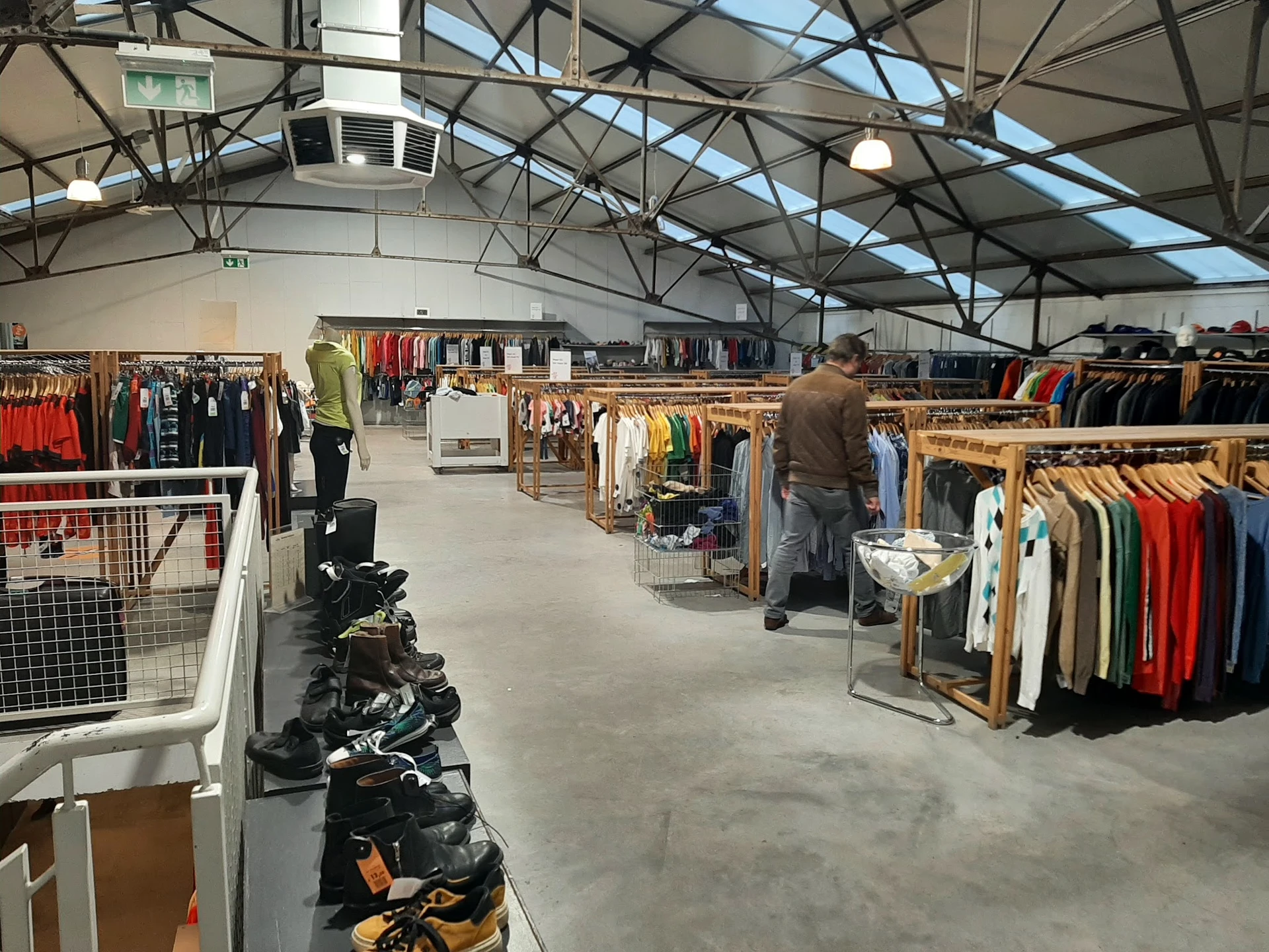 Ruime magazijnhal met rekken vol tweedehands kleding in verschillende kleuren, persoon aan het winkelen