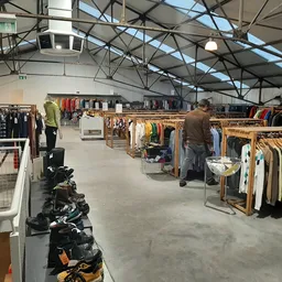 Ruime magazijnhal met rekken vol tweedehands kleding in verschillende kleuren, persoon aan het winkelen