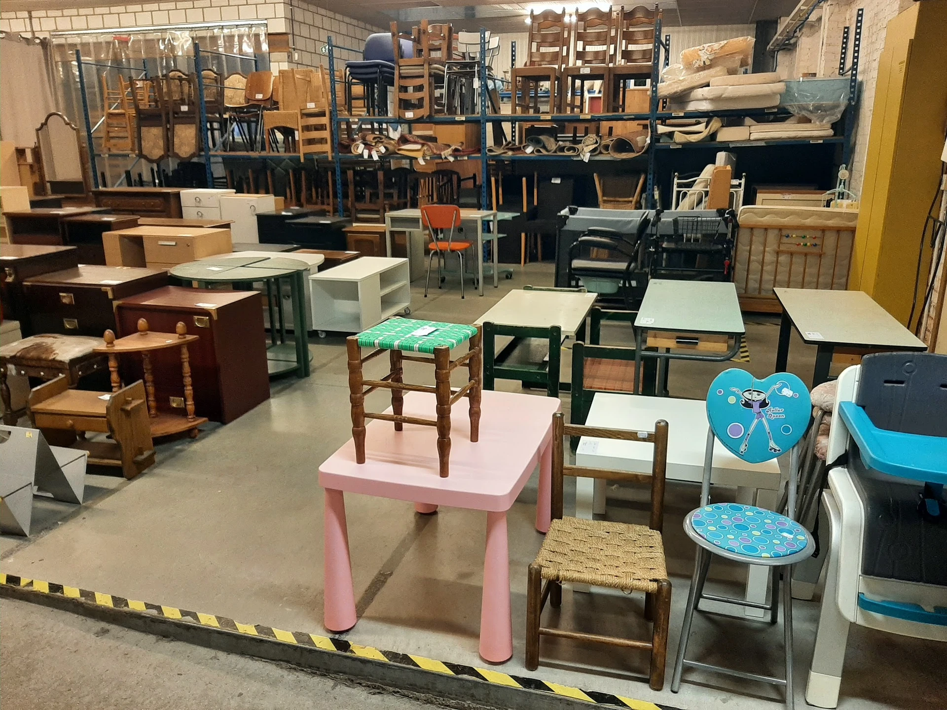 Diverse meubels en stoelen uitgestald in kringloopwinkel, waaronder roze tafel, houten stoelen en kinderstoelen