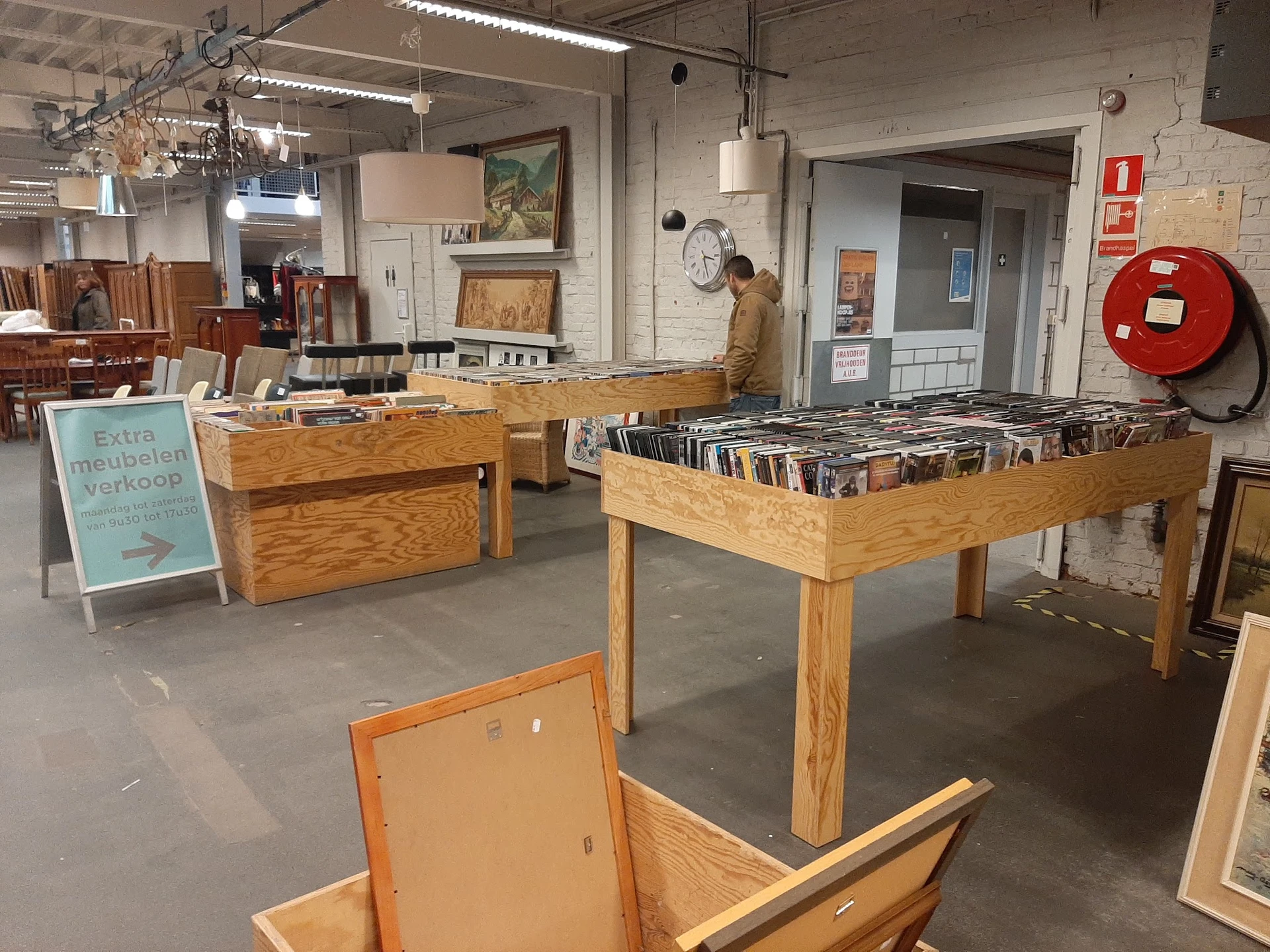 Winkelinterieur met houten tafels vol cd's en dvd's, stellingen op de achtergrond, rode brandsluscabinet aan de wand