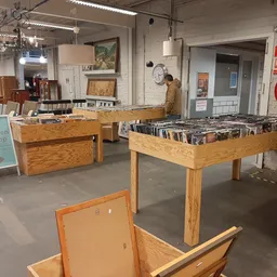 Winkelinterieur met houten tafels vol cd's en dvd's, stellingen op de achtergrond, rode brandsluscabinet aan de wand