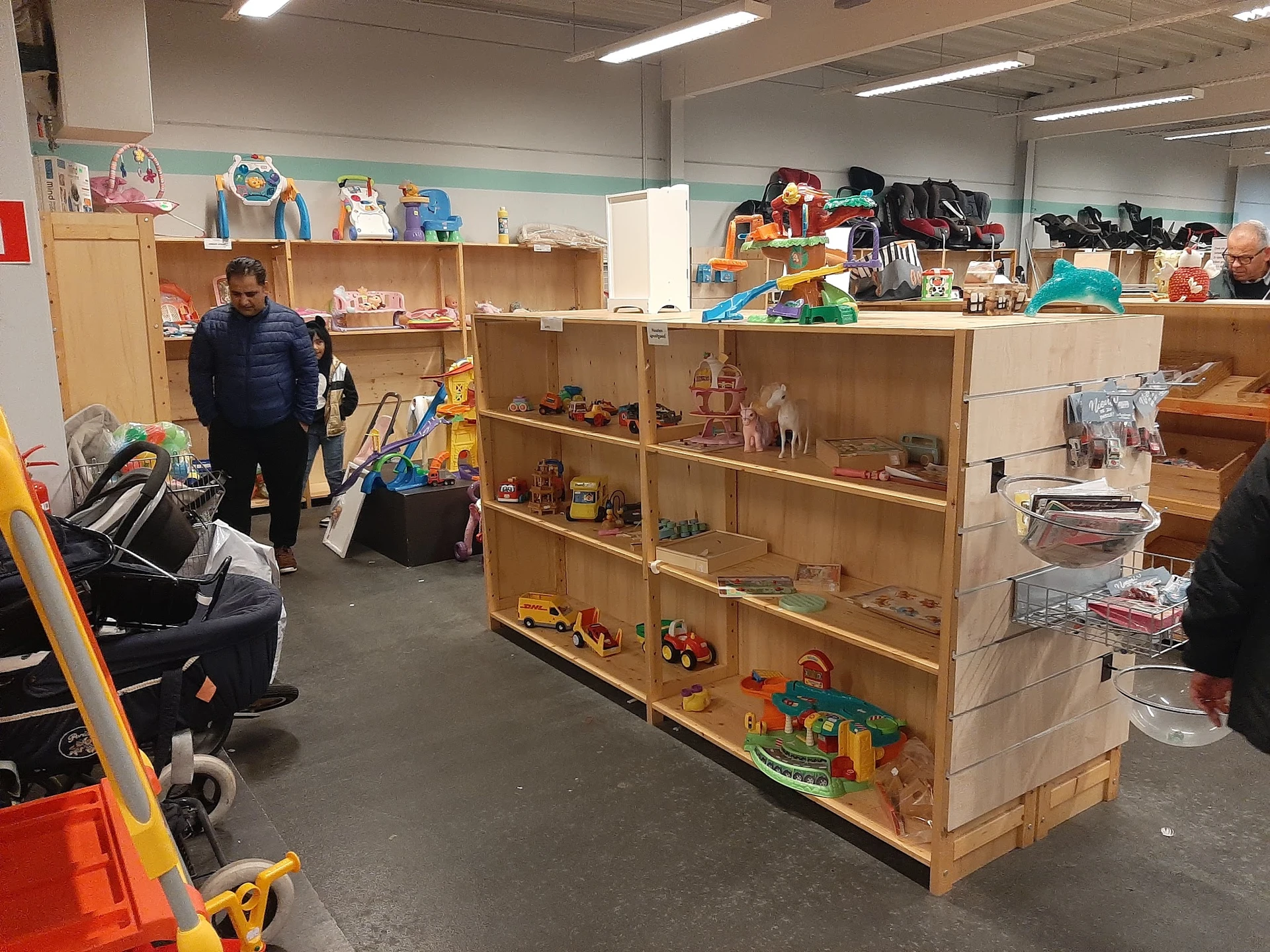 Winkelvloer met houten stellingen vol speelgoed, poppen en huishoudartikelen; twee bezoekers bladeren door spullen