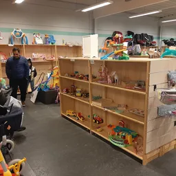 Winkelvloer met houten stellingen vol speelgoed, poppen en huishoudartikelen; twee bezoekers bladeren door spullen