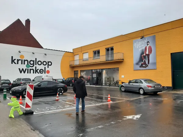 Gele gevel van kringloopwinkel met winkelnaam, parkeerplaats en personen voor de ingang
