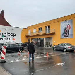 Gele gevel van kringloopwinkel met winkelnaam, parkeerplaats en personen voor de ingang
