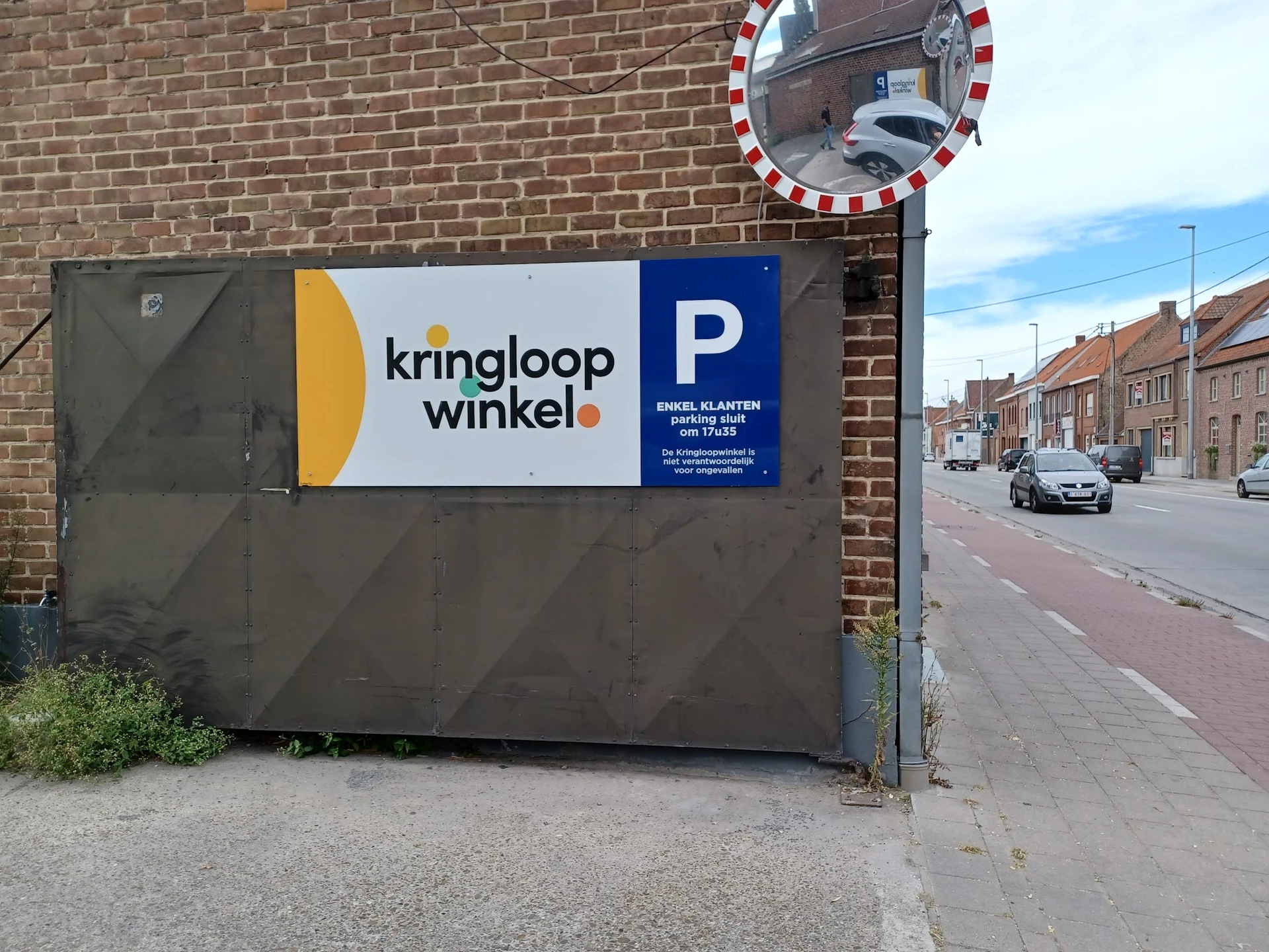 Grijze gevel van kringloopwinkel met logo en parkeerbordje, aan bakstenen muur langs straat