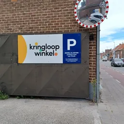 Grijze gevel van kringloopwinkel met logo en parkeerbordje, aan bakstenen muur langs straat