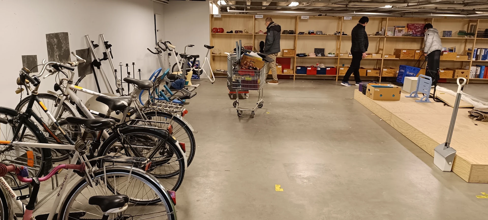 Winkelvloer met fietsen links, winkelwagen in het midden en houten stellingen met spullen op de achtergrond, enkele klanten i
