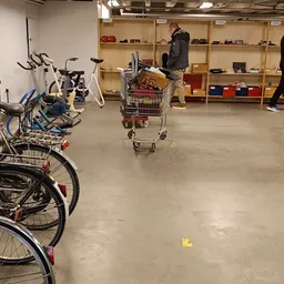 Winkelvloer met fietsen links, winkelwagen in het midden en houten stellingen met spullen op de achtergrond, enkele klanten i
