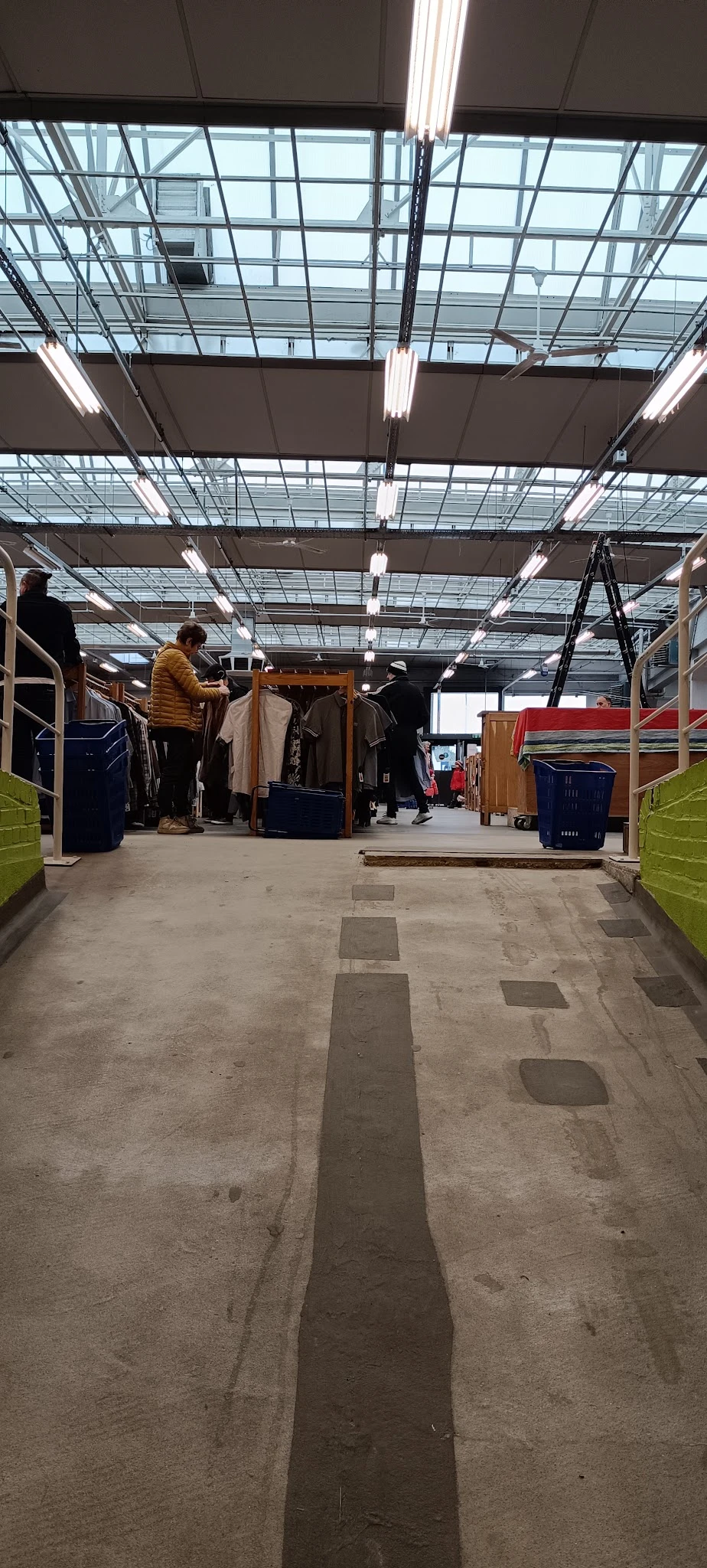 Winkelinterieur met kledingrekken, blauwe en groene plastic kratten, en klanten die kleding uitzoeken onder fluorescente verl
