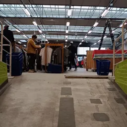 Winkelinterieur met kledingrekken, blauwe en groene plastic kratten, en klanten die kleding uitzoeken onder fluorescente verl