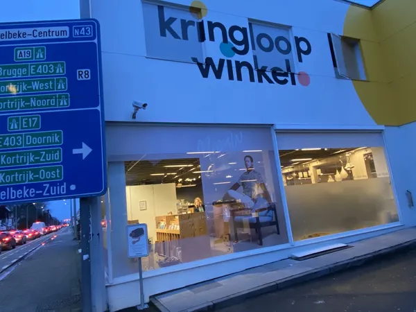 Blauwe gevel van Kringloop Winkel met verlichte etalageruiten aan avond, naast verkeersbord