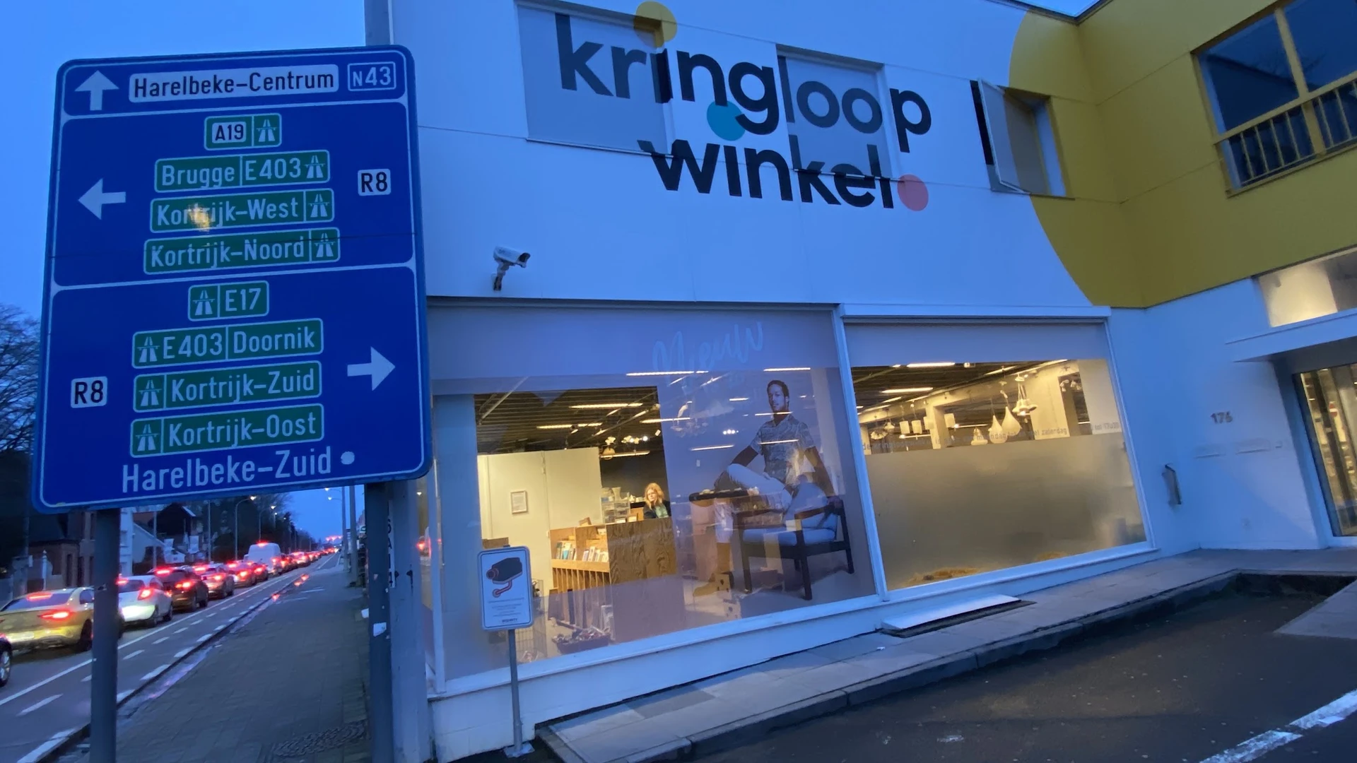 Blauwe gevel van Kringloop Winkel met verlichte etalageruiten aan avond, naast verkeersbord