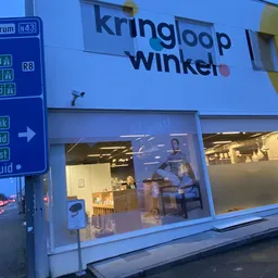 Blauwe gevel van Kringloop Winkel met verlichte etalageruiten aan avond, naast verkeersbord