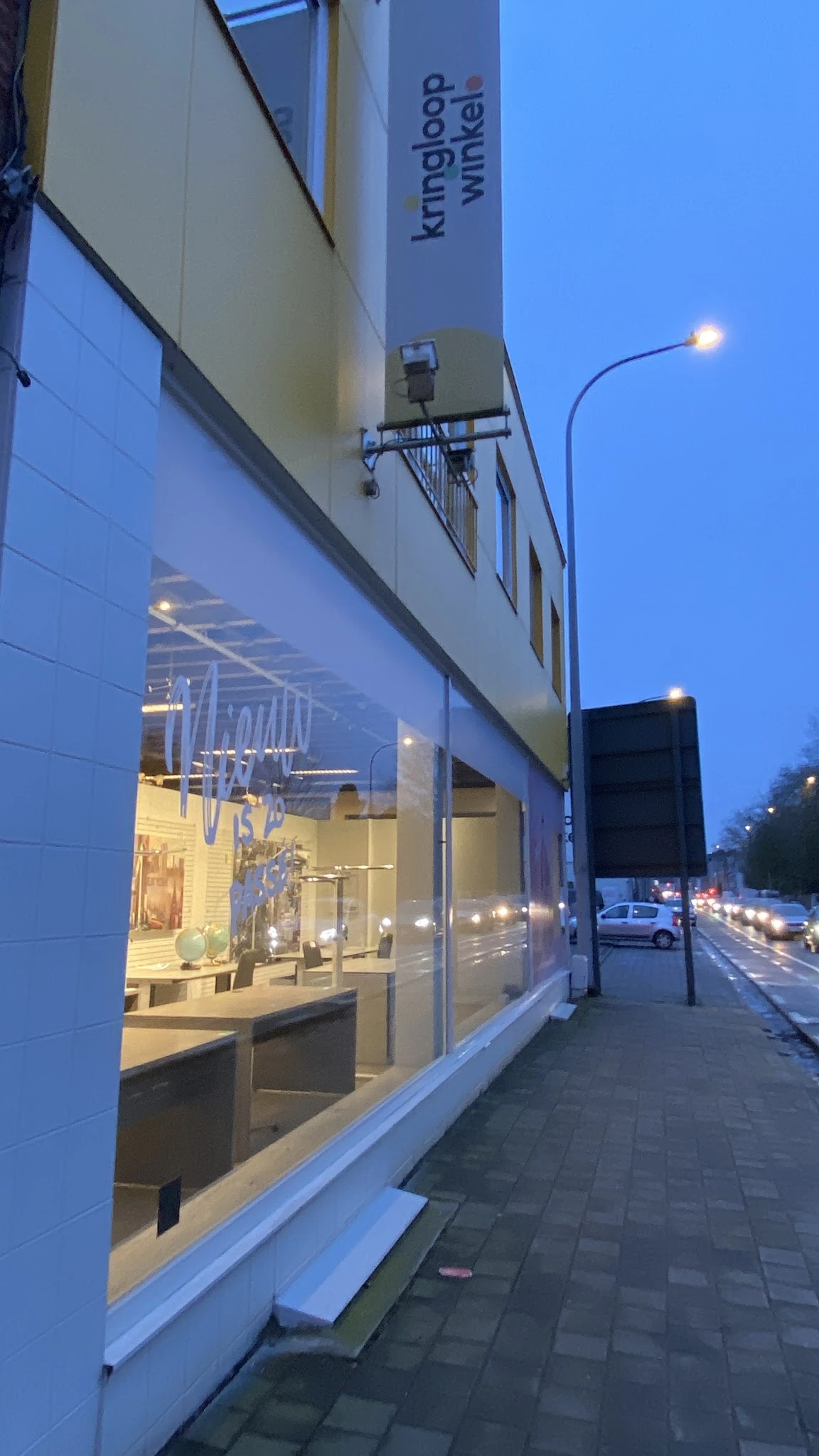Gevelfront van kringloopwinkel met verlichte etalage aan schemering, winkelramen en ingang zichtbaar