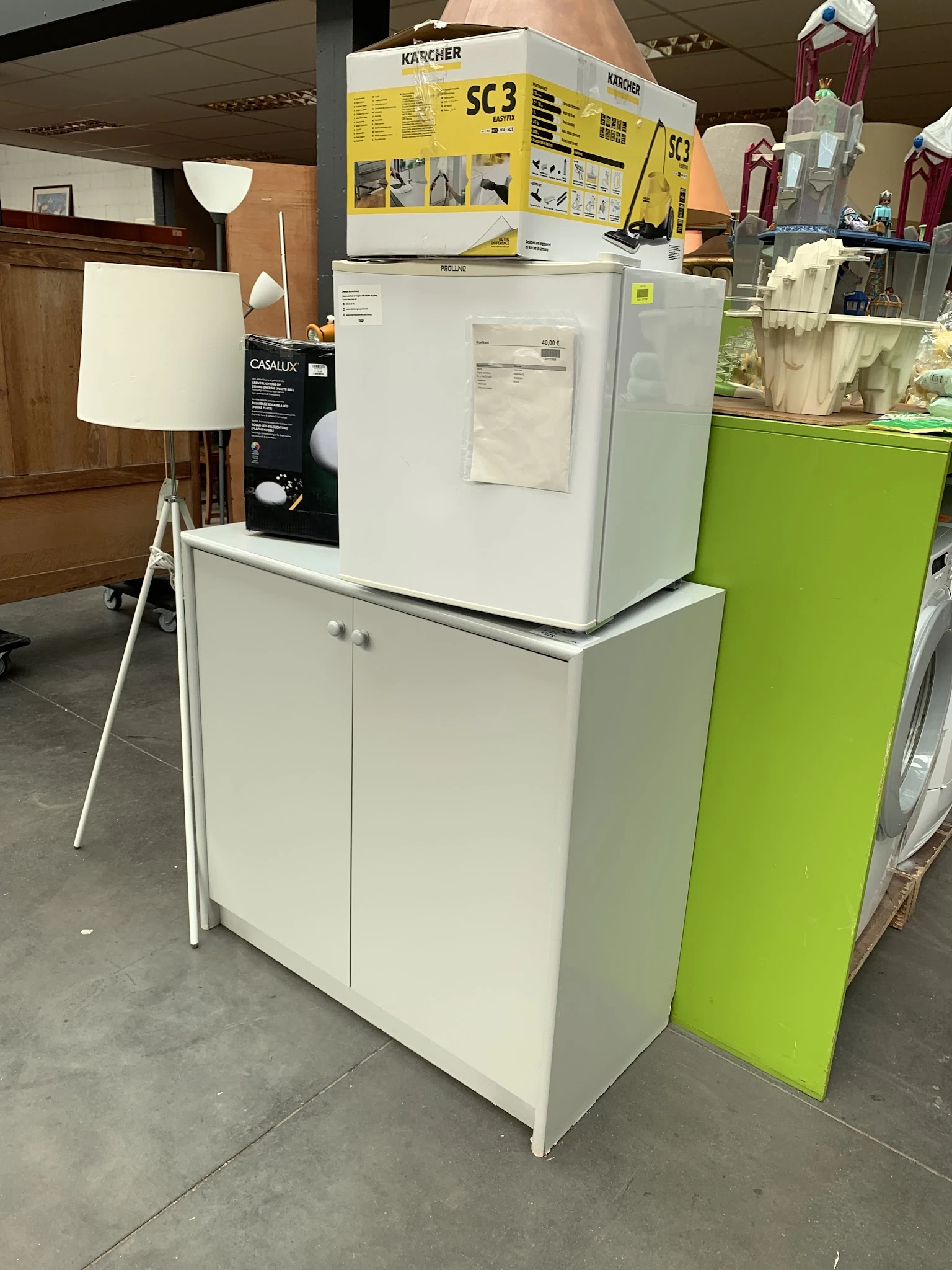 Uitgestalde meubels in de winkel: witte kast met deuren, witte lamp en groene kasten op de achtergrond