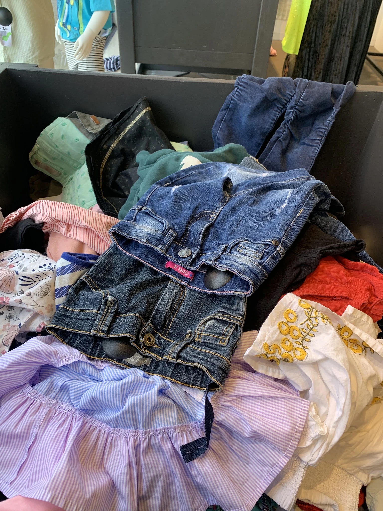 Stapel tweedehands kleding met jeans, shirts en andere kledingstukken in verschillende kleuren