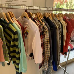 Rekken met tweedehands kleding op hangers, waaronder gestreepte truien, shirts met prints, geruite blouses en jurkjes in vers