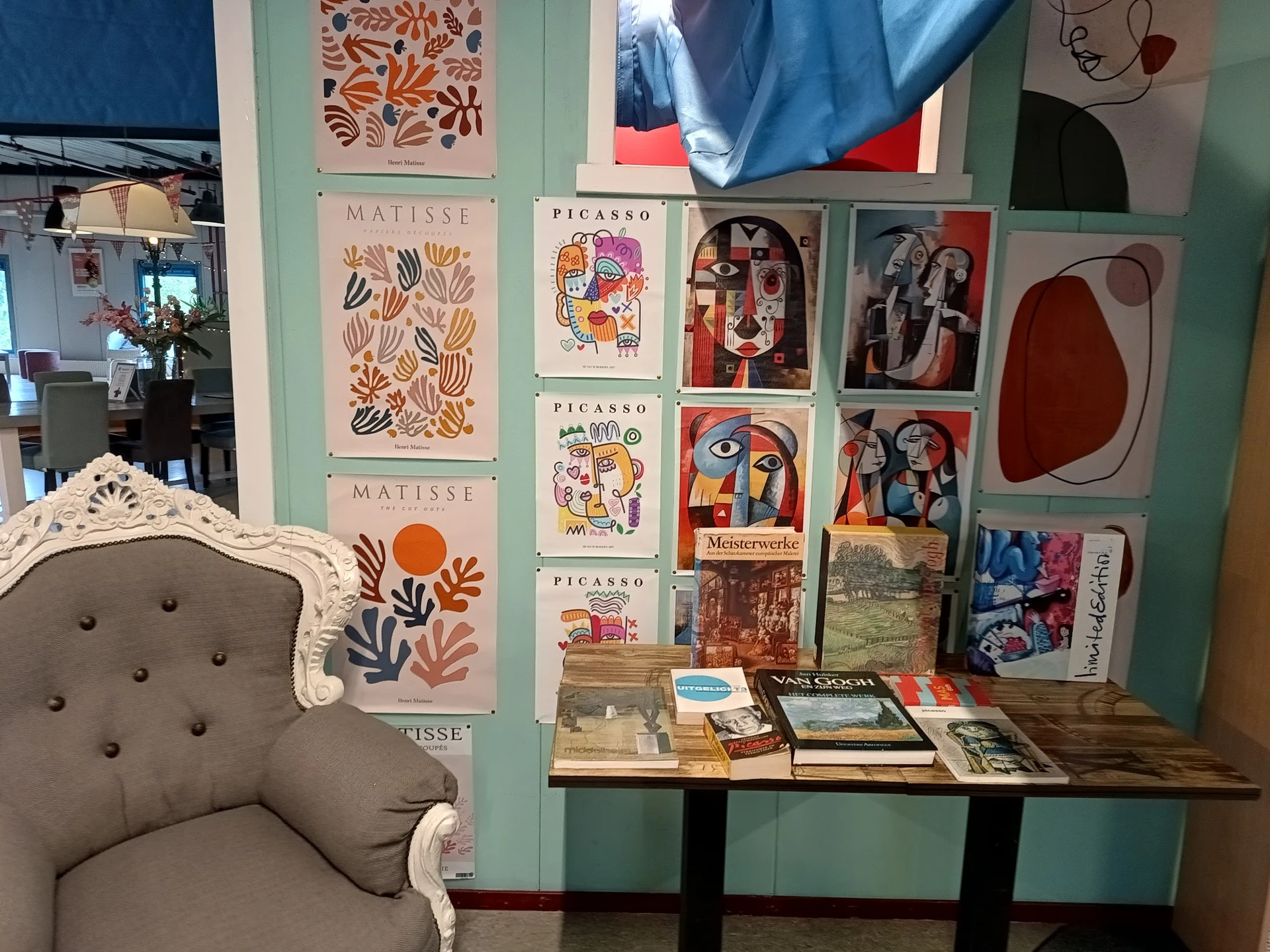 Wand met kunstafdrukken van Matisse en Picasso, fotolijsten, boeken en decoratieve items op een houten tafel, naast een vinta