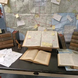 Tafel met opengeslagen antieke boeken, kaarten en notities in een winkelinterieur met boekenrekken op de achtergrond