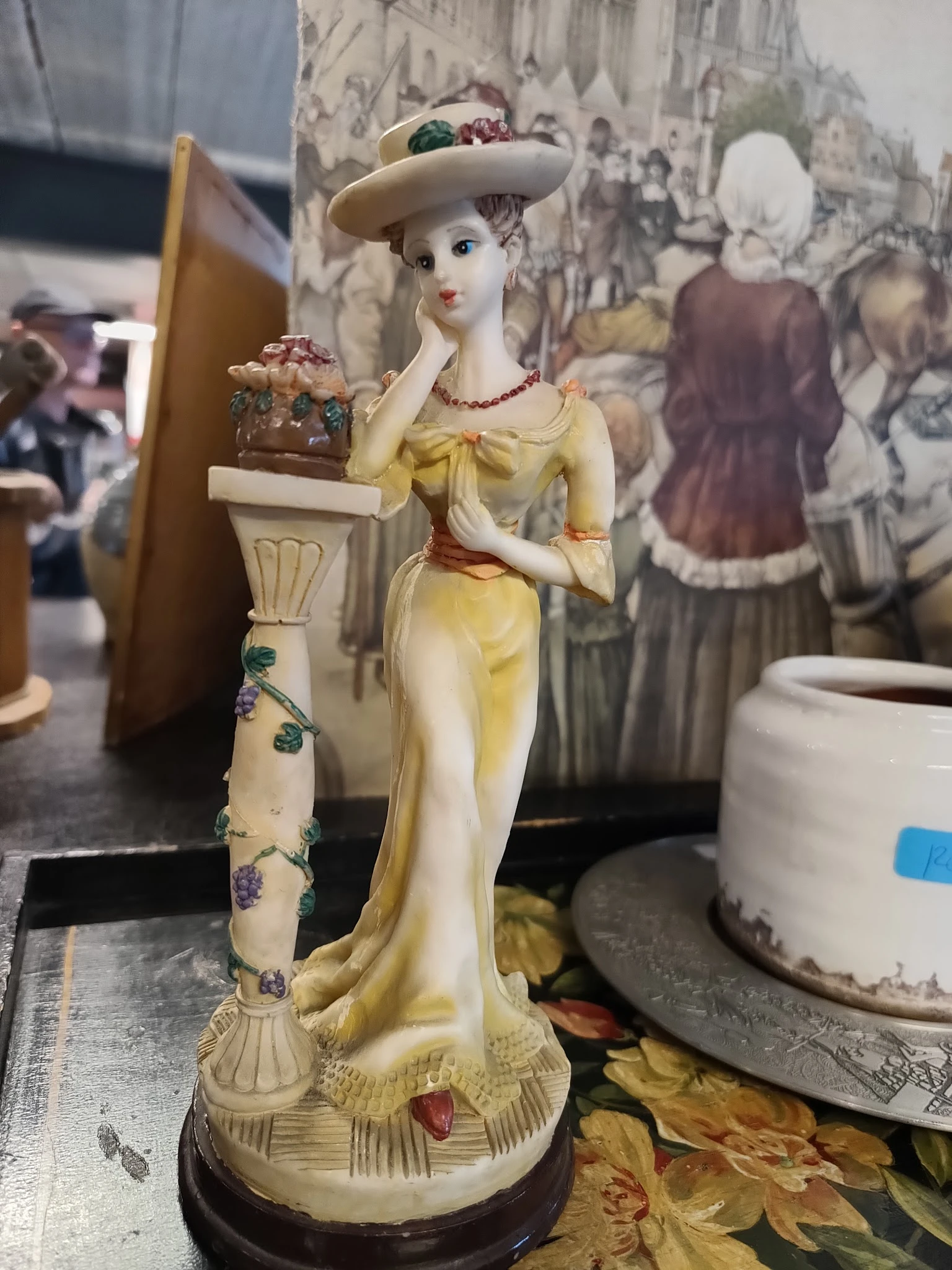 Close-up van een geel porselein beeldje van een vrouw in klassieke stijl, met rode details en een decoratieve zuil naast zich