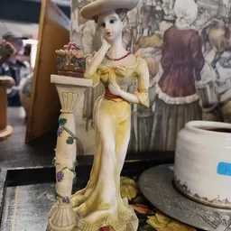 Close-up van een geel porselein beeldje van een vrouw in klassieke stijl, met rode details en een decoratieve zuil naast zich