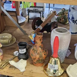 Diverse gebruikte huishoudartikelen op een houten tafel: servies, decoratiefiguren, vazen, een bel en ander brocante