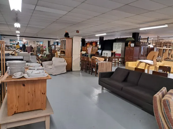 Ruim winkelinterieur met diverse meubels, een zwarte bank, houten kasten, stoelen en huishoudelijke artikelen op tafels