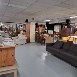 Ruim winkelinterieur met diverse meubels, een zwarte bank, houten kasten, stoelen en huishoudelijke artikelen op tafels