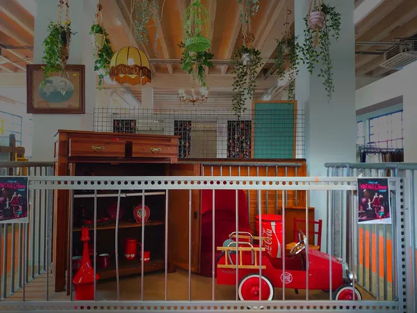 Speelhoek in kringloopwinkel met rode vintage speelgoedauto's achter hek, hangplanten aan plafond