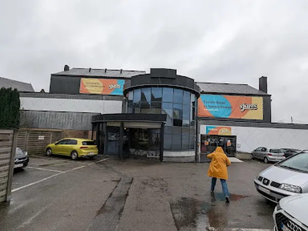 Exterieur van kringloopwinkel met ronde ingang, gele auto op parkeerplaats en persoon in geel jack