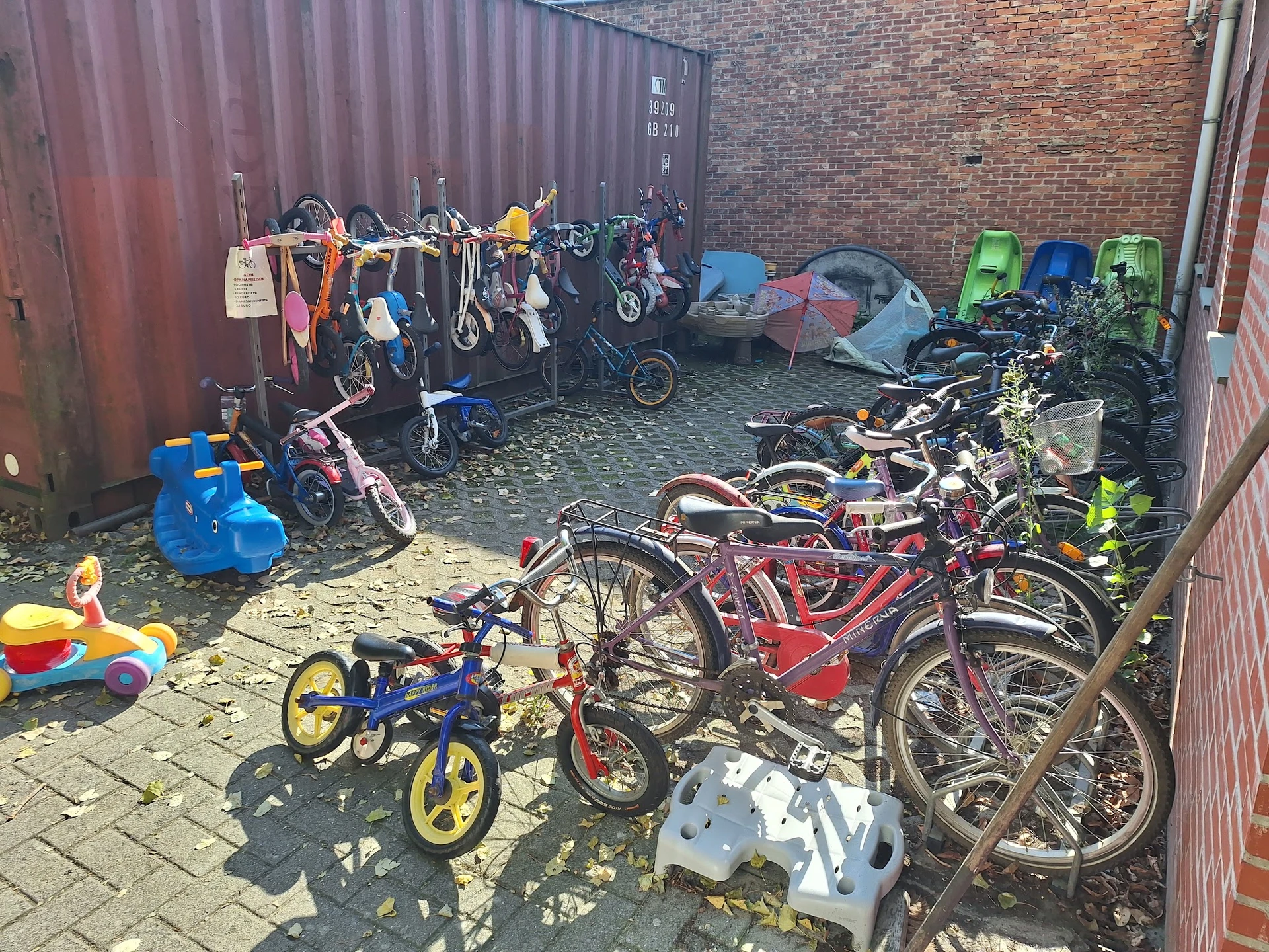 Verzameling tweedehands fietsen, kinderfietsjes en speelgoed in een opslagruimte tegen een bakstenen muur